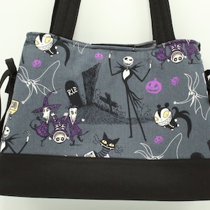Op de afbeelding: Een grijze tote bag met een zwarte bodem en zwarte hengsels. De tas heeft een print van personages uit de film Nightmare Before Christmas, waaronder Jack Skellington, Sally en Zero. De print bevat ook pompoenen en een grafsteen met de inscriptie "RIP".