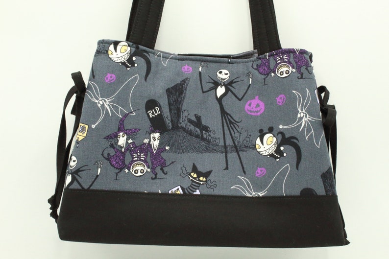 Op de afbeelding: Een grijze tote bag met een zwarte bodem en zwarte hengsels. De tas heeft een print van personages uit de film Nightmare Before Christmas, waaronder Jack Skellington, Sally en Zero. De print bevat ook pompoenen en een grafsteen met de inscriptie "RIP".