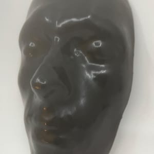 May include: A dark, glossy, human face mask with a smooth surface. The mask is a solid color, with a slightly reflective sheen. The features are clearly defined, including the nose, eyes, and mouth. The mask is likely made of a hard material.