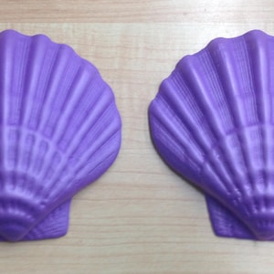 Puede incluir: Dos jaboneras de color morado con forma de concha marina. Las jaboneras están hechas de un material liso y brillante y tienen un diseño detallado y realista de concha marina.