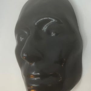 May include: A dark, glossy black mask of a human face. The mask has a smooth surface with subtle details of the nose, lips, and eyes. The mask is likely for decorative or theatrical purposes.