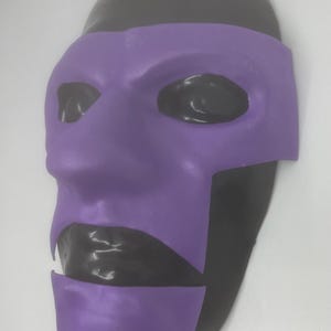 Peut inclure: Un masque bicolore avec un couvre-visage violet et une tête noire. La section violette comprend les zones des yeux et de la bouche. Le masque est probablement destiné à un costume ou à un usage théâtral.