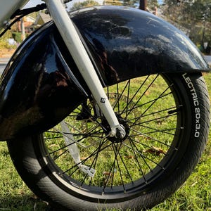 Pu&ograve; includere: Primo piano di un parafango e di uno pneumatico per bicicletta neri. Lo pneumatico riporta la scritta "LECTRIC 20x3.0" in bianco. La forcella della bicicletta &egrave; bianca. Lo sfondo &egrave; erba verde.