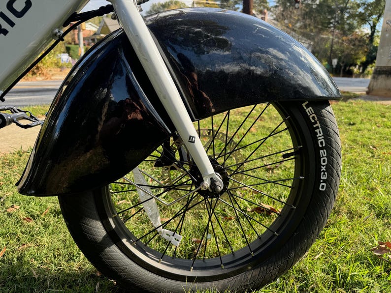 Pu&ograve; includere: Primo piano di un parafango e di uno pneumatico per bicicletta neri. Lo pneumatico riporta la scritta "LECTRIC 20x3.0" in bianco. La forcella della bicicletta &egrave; bianca. Lo sfondo &egrave; erba verde.