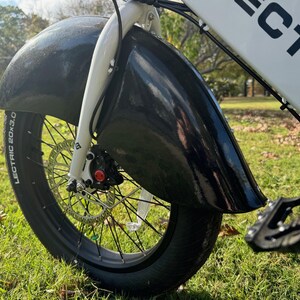 Pu&ograve; includere: Primo piano di un parafango e pneumatico per bicicletta elettrica neri. Lo pneumatico &egrave; nero con la scritta "LECTRIC 20x3.0". La bici ha forcella e telaio bianchi. Lo sfondo &egrave; erba verde.