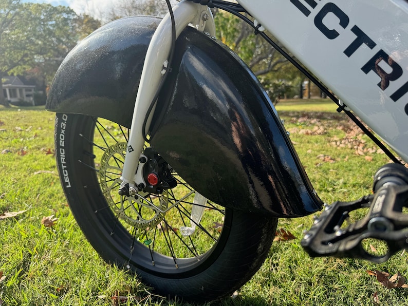 Pu&ograve; includere: Primo piano di un parafango e pneumatico per bicicletta elettrica neri. Lo pneumatico &egrave; nero con la scritta "LECTRIC 20x3.0". La bici ha forcella e telaio bianchi. Lo sfondo &egrave; erba verde.