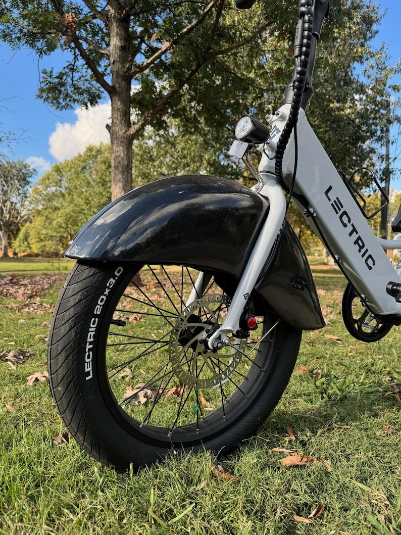 Pu&ograve; includere: Primo piano di una e-bike Lectric con parafango anteriore e pneumatico neri. Lo pneumatico riporta la scritta "LECTRIC 20x3.0". La bici &egrave; bianca e grigia con faro e freni a disco. Lo sfondo &egrave; erba verde e alberi.