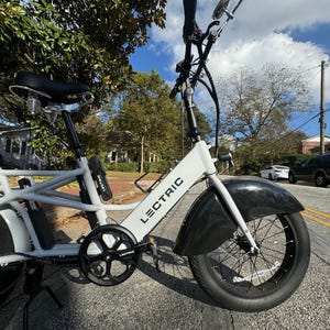 Pu&ograve; includere: E-bike cargo Lectric bianca con parafanghi e pneumatici neri. La bici ha sella, pedali e batteria neri. Il telaio &egrave; bianco con la scritta "LECTRIC" stampata. La bici &egrave; parcheggiata su una strada asfaltata.