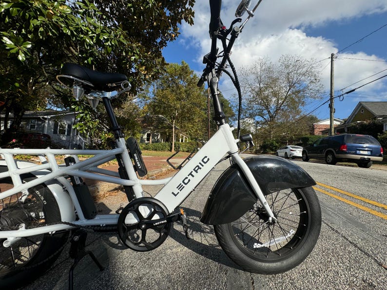 Pu&ograve; includere: E-bike cargo Lectric bianca con parafanghi e pneumatici neri. La bici ha sella, pedali e batteria neri. Il telaio &egrave; bianco con la scritta "LECTRIC" stampata. La bici &egrave; parcheggiata su una strada asfaltata.