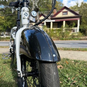 Pu&ograve; includere: Primo piano di una bicicletta elettrica in bianco e nero. Lo pneumatico anteriore e il parafango nero sono a fuoco, con un telaio bianco e un faro visibile. Lo sfondo mostra una strada residenziale con case e alberi.