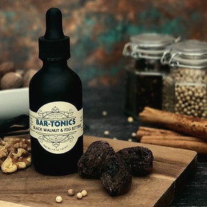 May include: A black glass bottle with a dropper top and a label that reads "BAR-TONICS BLACK WALNUT & FIG BITTERS" with a net weight of 2 fl oz (30 ml). The bottle is sitting on a wooden cutting board with dried figs, walnuts, and white peppercorns.