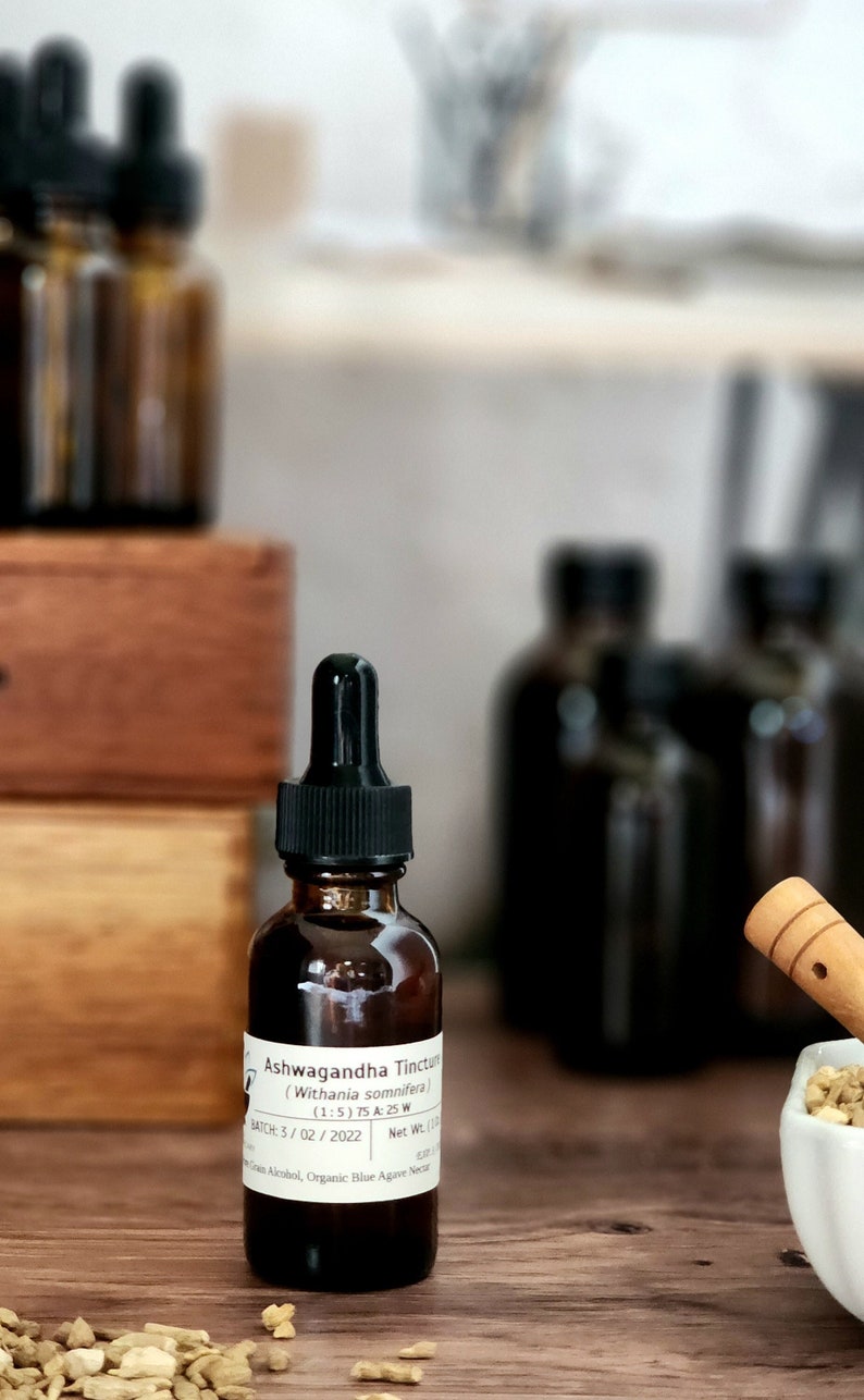 May include: A brown glass bottle with a black dropper lid and a white label that reads "Ashwagandha Tincture (Withania somnifera) (1:5) 75 A:25 W BATCH: 3/02/2022 Net Wt. 1 fl oz" The bottle is sitting on a wooden surface with a small white bowl filled with dried herbs and a wooden spoon.