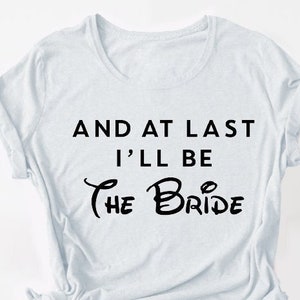 Puede incluir: Camiseta blanca con texto negro que dice "And at last I'll be the Bride" con una fuente Disney estilizada para "Bride".