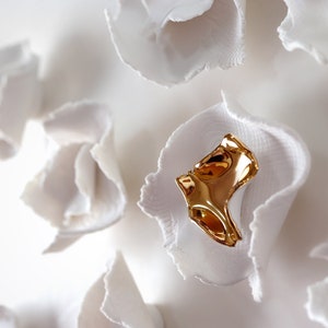 May include: A gold-colored metal piece of jewelry with a unique, abstract design. The jewelry is displayed on a white background.
