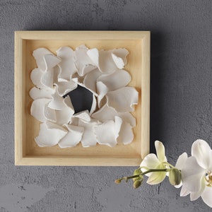 May include: A wooden frame with a white, organic, flower-like design inside. The design is made of many small, white pieces that are arranged in a circular pattern. The frame is on a gray background.