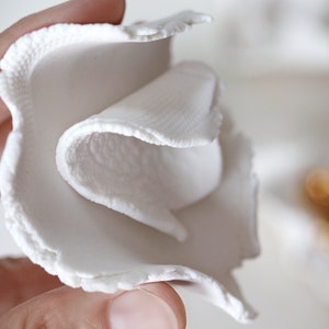 May include: A white ceramic flower with a textured surface. The flower is folded over itself, creating a layered effect.