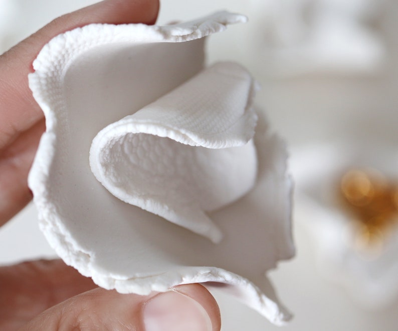 May include: A white ceramic flower with a textured surface. The flower is folded over itself, creating a layered effect.