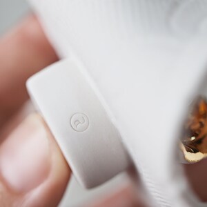 May include: Close-up of a white ceramic object with a gold accent. The object has a small, round, raised detail with the letter "R" inside.