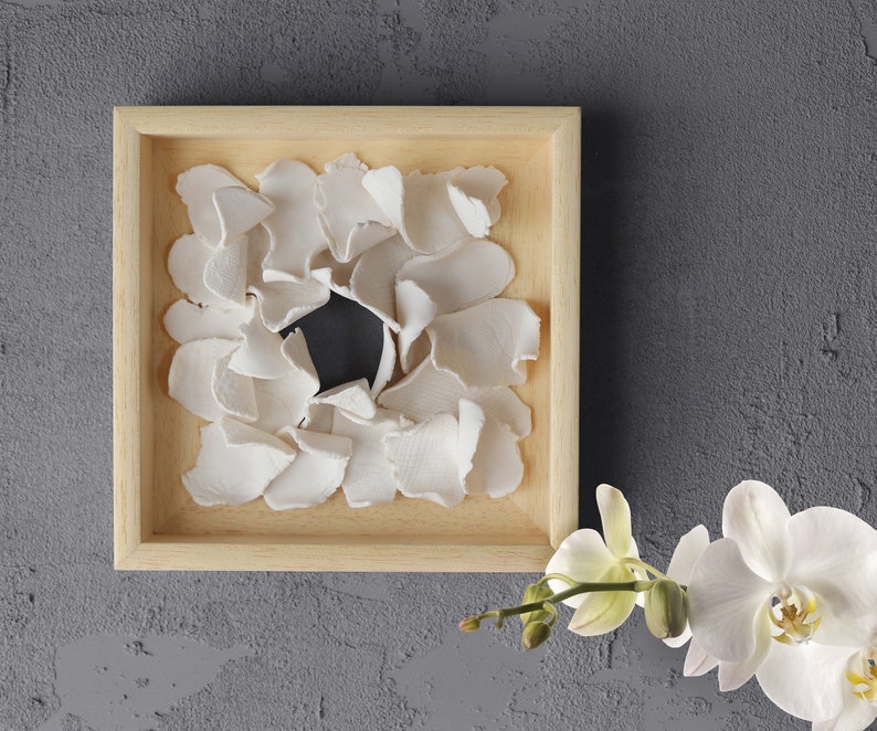 May include: A wooden frame with a white, organic, flower-like design inside. The design is made of many small, white pieces that are arranged in a circular pattern. The frame is on a gray background.