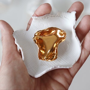 May include: A gold-colored, abstract, organic-shaped sculpture sits on a white ceramic dish. The dish is held in a person's hand.