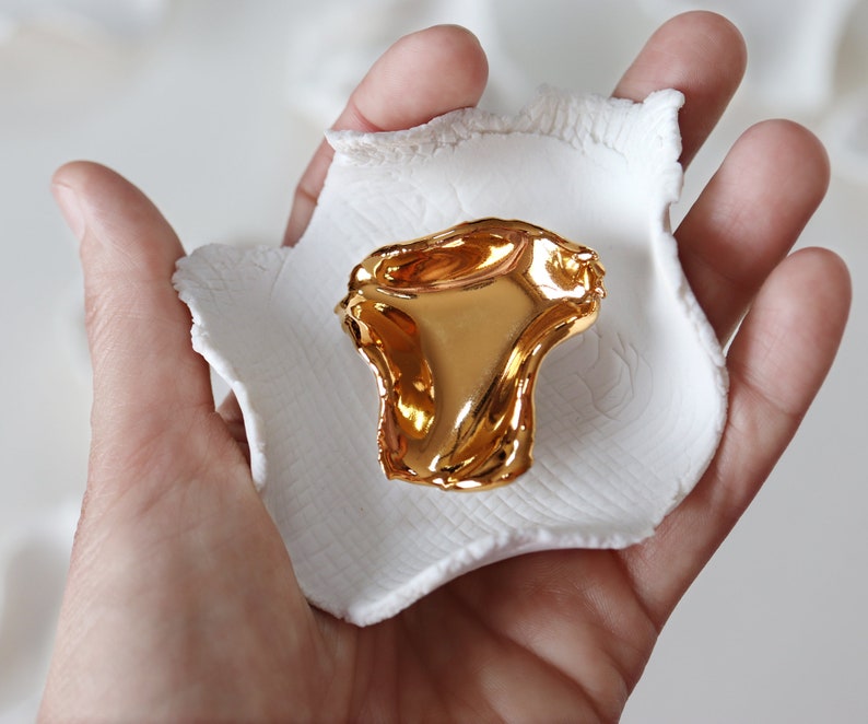 May include: A gold-colored, abstract, organic-shaped sculpture sits on a white ceramic dish. The dish is held in a person's hand.
