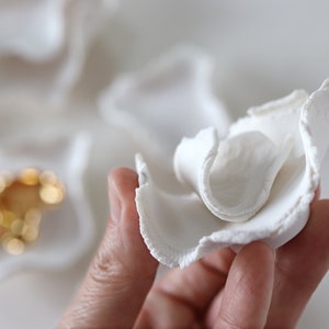 May include: A white ceramic flower with a ruffled texture. The flower is held in a hand, and the background is a white surface.