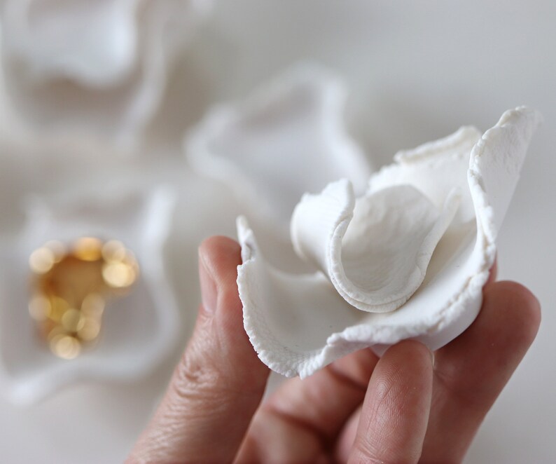May include: A white ceramic flower with a ruffled texture. The flower is held in a hand, and the background is a white surface.