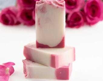 May include: Three stacked bars of handmade soap with a white and pink swirl design. The soap has a rectangular shape with rounded edges. Pink rose petals are scattered on the surface, and pink roses are in the background.