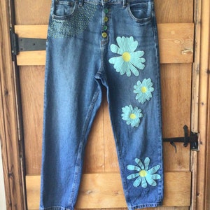 May include: Blue denim jeans with embroidered flowers in shades of blue and green. The flowers are arranged on the front of the jeans, with one on each leg and two on the front.