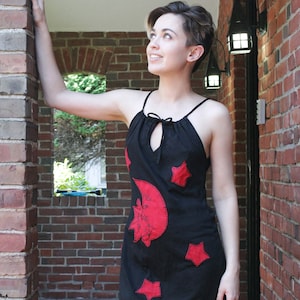 May include: A woman wearing a black sleeveless dress with a red crescent moon and stars design. The dress has a keyhole opening at the neckline and a tie closure.