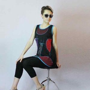 May include: A person wearing a black tunic with a colorful abstract design, black leggings, and sandals with a silver strap. The tunic has a blue trim and red accents. The person is wearing sunglasses and sitting on a chrome stool.