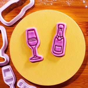 Può includere: Tagliapasta e biscotti a tema champagne. L'immagine mostra una bottiglia di champagne e un biscotto a forma di calice di champagne, entrambi con glassa rosa, su un piatto giallo. Sono visibili anche diversi tagliapasta in plastica bianca.