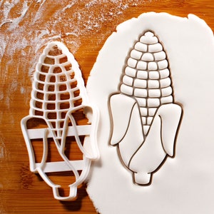 May include: A white plastic cookie cutter in the shape of an ear of corn. The cutter is shown on a wooden surface with a white background.