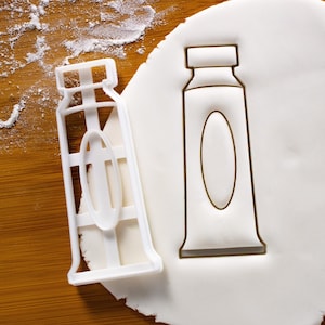 May include: Two white plastic cookie cutters shaped like tubes of toothpaste. The cutters are on a white surface.