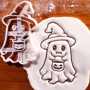 Peut inclure: Emporte-pièce blanc en forme de fantôme d'Halloween portant un chapeau de sorcière et tenant un seau de citrouille. Le fantôme est pressé dans une pâte blanche, créant un motif en relief. Le découpoir est à côté du fantôme en relief.