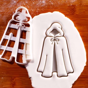 May include: A white plastic cookie cutter in the shape of a hooded cloak with a bow at the neck. The cutter is next to a piece of white dough with the cloak shape pressed into it. The background is a wooden surface.