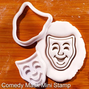 May include: A white plastic comedy mask cookie cutter and a matching mini stamp. The stamp is pressed into a circular piece of white dough, creating a detailed impression of a smiling theatrical face. The text Comedy Mask Mini Stamp is visible.