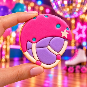 May include: A round cookie decorated with royal icing to resemble a roller skating helmet. The design features a bright pink upper section with small teal dots and a white star, and a lavender lower section with white straps and pink accents.