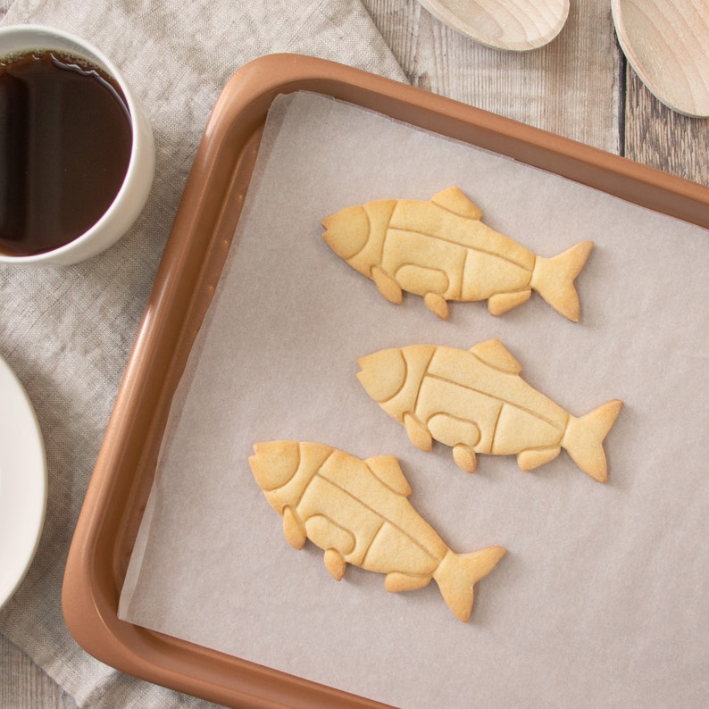 Puede incluir: Tres galletas con forma de pez sobre papel de hornear dentro de una bandeja de horno de color cobre. Una taza de l&iacute;quido oscuro y cucharas de madera son visibles en el fondo. Las galletas son de color marr&oacute;n claro con dise&ntilde;os recortados.