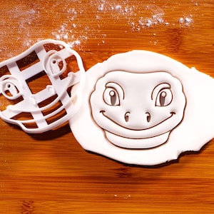 May include: A white plastic cookie cutter shaped like a cartoon lizard face with large eyes and a wide smile, positioned next to a piece of white rolled dough that has been imprinted with the same lizard face design, showing clear facial features.