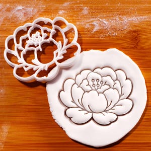 May include: A white plastic peony flower-shaped cookie cutter next to a peony-shaped imprint in white dough. The cutter is on a wooden surface with a dusting of white powder.