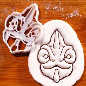 May include: A white plastic cookie cutter shaped like a chameleon head, featuring a pointed snout, large circular eyes, and a curved mouth. Beside it, a piece of white dough shows the embossed impression of the same chameleon face design.