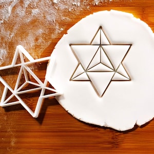 May include: A white plastic cookie cutter shaped like a six-pointed star with internal geometric lines, positioned next to a circular piece of white rolled dough that has been imprinted with the same star design, showing clear triangular segments.