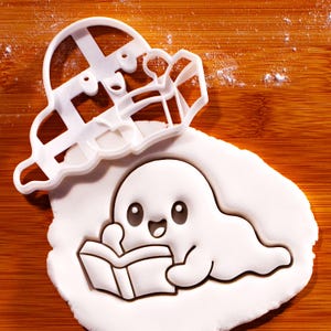 Op de afbeelding: Een witte plastic koekjesvorm in de vorm van een geest die een boek leest. De vorm ligt boven een stuk wit uitgerold deeg dat is bedrukt met hetzelfde geestfiguurtje, met een lachend gezicht en een open boek dat door handjes wordt vastgehouden.