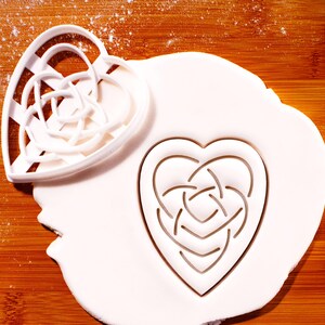 May include: A white plastic heart-shaped cookie cutter with a Celtic knot design, next to a heart-shaped cutout in white dough. The cutter is on a wooden surface, with a dusting of white powder.