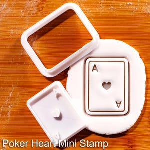 以下が含まれることがあります： 白いポーカーをテーマにしたミニスタンプセット。正方形のフレーム、ハートのエースカードのスタンプ、ハートの別のスタンプが含まれています。「Poker Heart Mini Stamp」というテキストが画像の下部に表示されています。