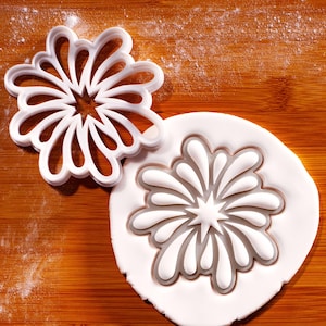 May include: A white plastic flower-shaped cookie cutter next to a flower-shaped imprint in white dough. The cutter has a star-shaped center with curved petal-like extensions. The dough imprint shows the same design, creating a floral pattern.