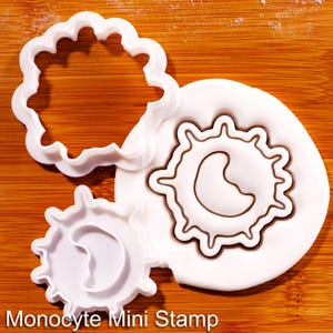 May include: A white plastic cookie cutter and matching stamp set shaped like a monocyte cell. The set includes a large outline cutter, a smaller cutter, and a stamped impression in white dough featuring the text Monocyte Mini Stamp.