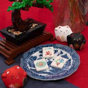 Puede incluir: Un bons&aacute;i en miniatura hecho de bloques marrones y verdes se encuentra sobre un soporte de madera. Un plato azul y blanco contiene tres baldosas decoradas. Tambi&eacute;n hay figuritas de gatos rojos, blancos y negros.