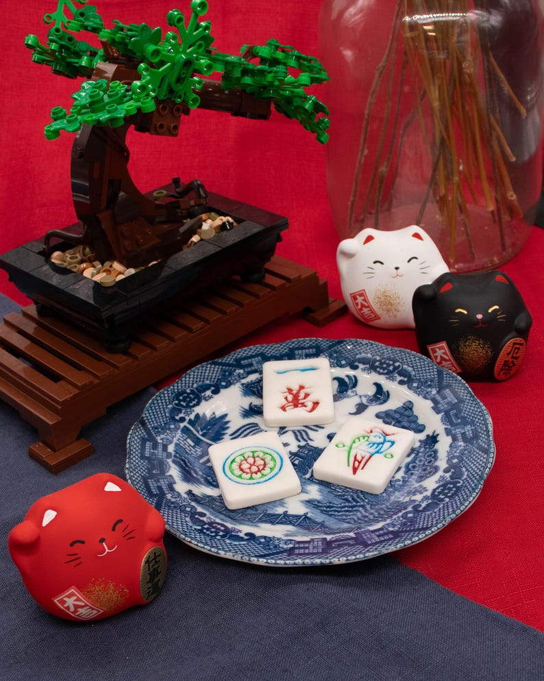 Puede incluir: Un bons&aacute;i en miniatura hecho de bloques marrones y verdes se encuentra sobre un soporte de madera. Un plato azul y blanco contiene tres baldosas decoradas. Tambi&eacute;n hay figuritas de gatos rojos, blancos y negros.